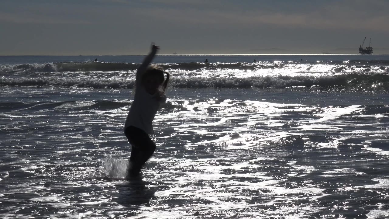 A silhouetted young girl in Huntington Beach lifts one arm high while splashing through shallow ocean water, with bright surf bands behind her.