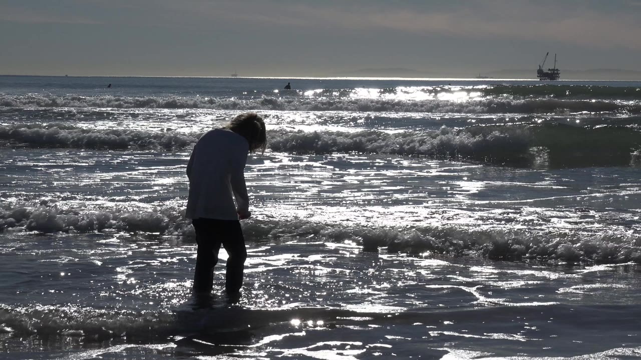 At Huntington Beach, California, a young girl stands in shallow water as backlit waves and wet sand reflect bright sun across the frame.
