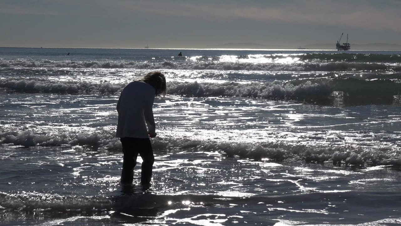 Child at the waterline facing bright waves and reflective wet sand.
