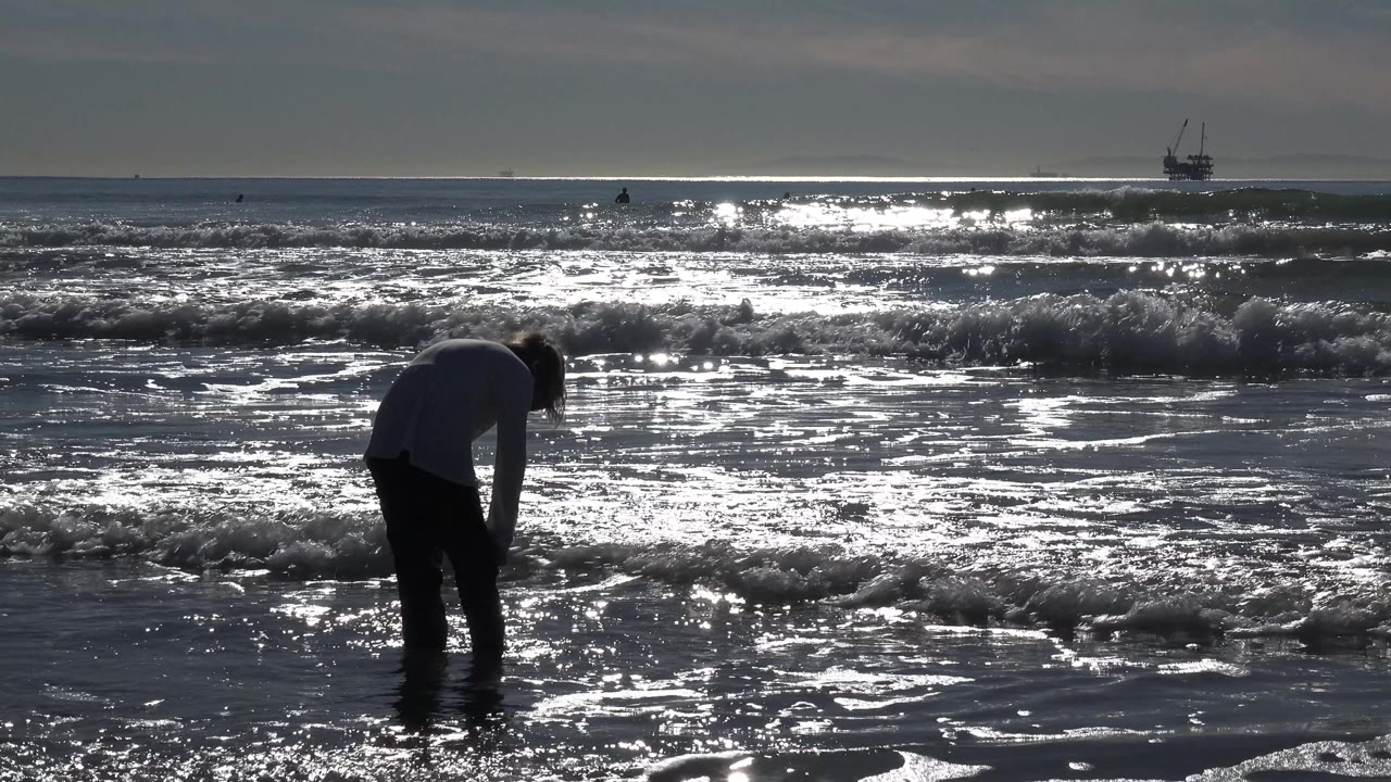 At Huntington Beach, California, the young girl bends forward in silhouette while foamy water spreads across the reflective sand beneath a bright ocean horizon.
