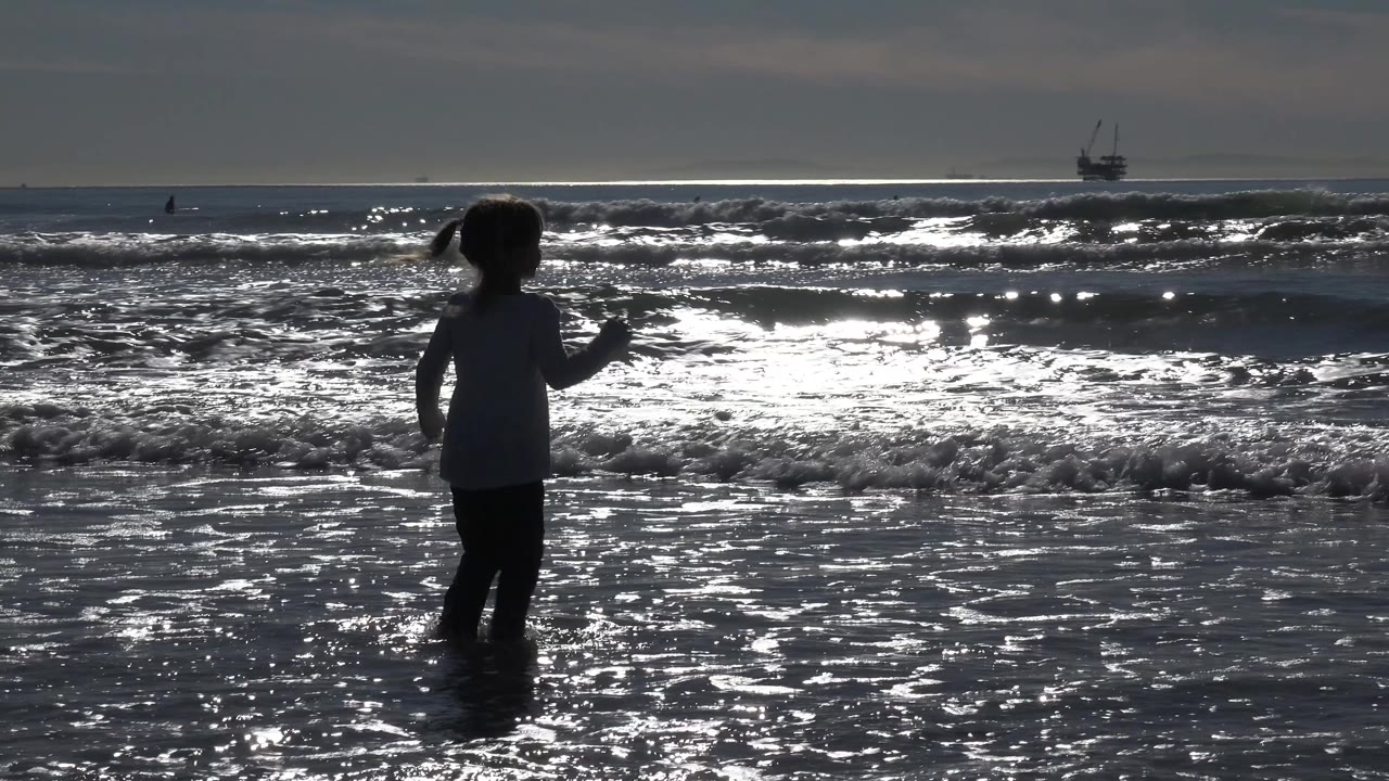 A child stands near the left side of the frame in shallow water at Huntington Beach, California, backlit by bright ocean waves and a distant vessel on the horizon.