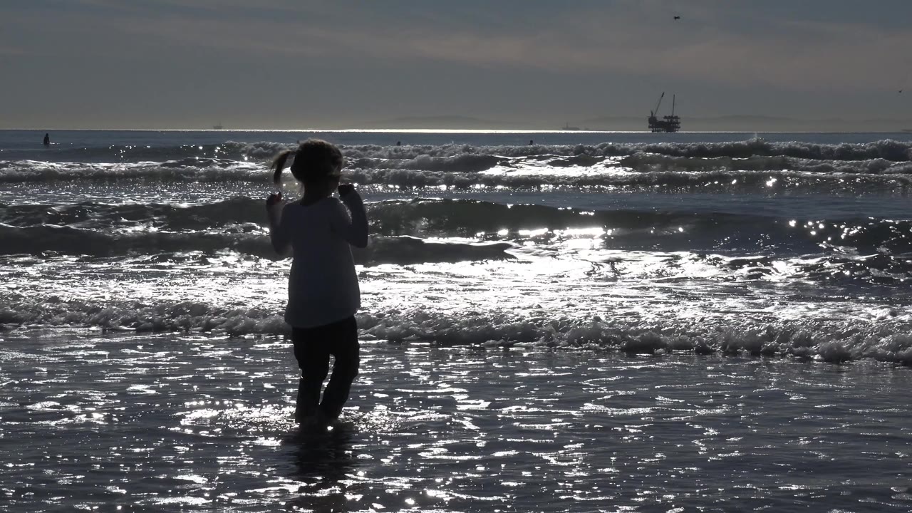 A child appears toward the right of the Huntington Beach frame as foamy waves roll across the midground and bright water reflects across the wet sand.