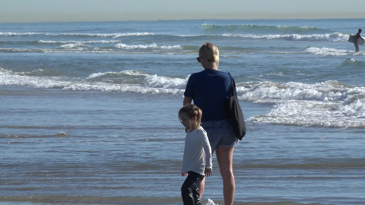 A woman and young child stand near shallow water at Huntington Beach, California, with breaking waves and a distant surfer in the background.