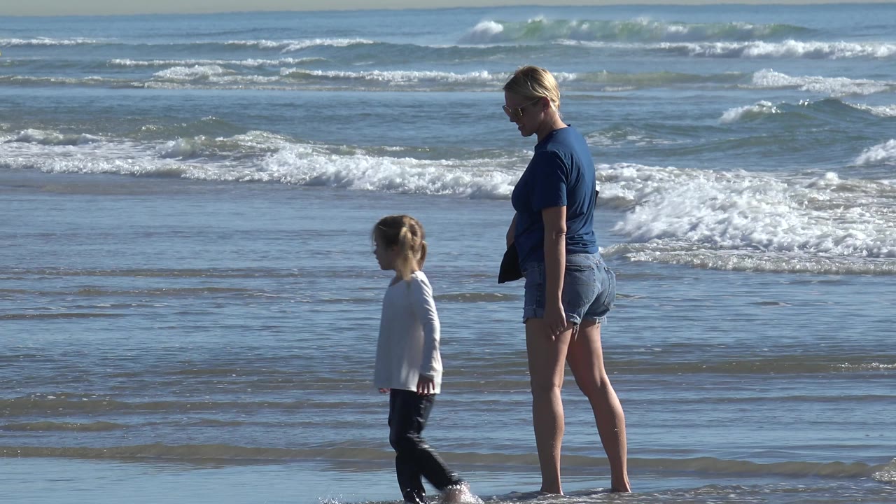 A woman and child walk along wet sand at Huntington Beach, California, with white surf behind them and the adult carrying a dark bag.