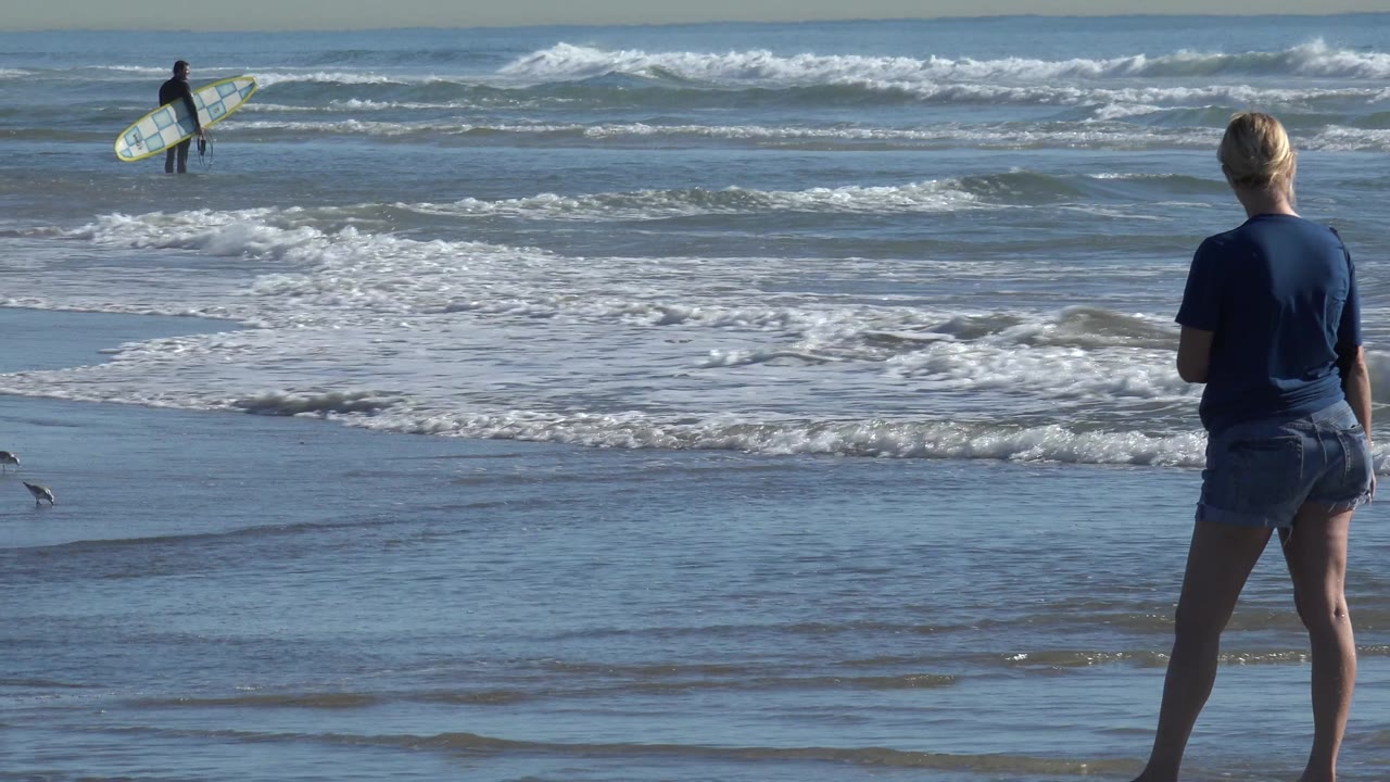 At Huntington Beach, a woman stands in the foreground facing the surf while a distant surfer carries a board near breaking waves.