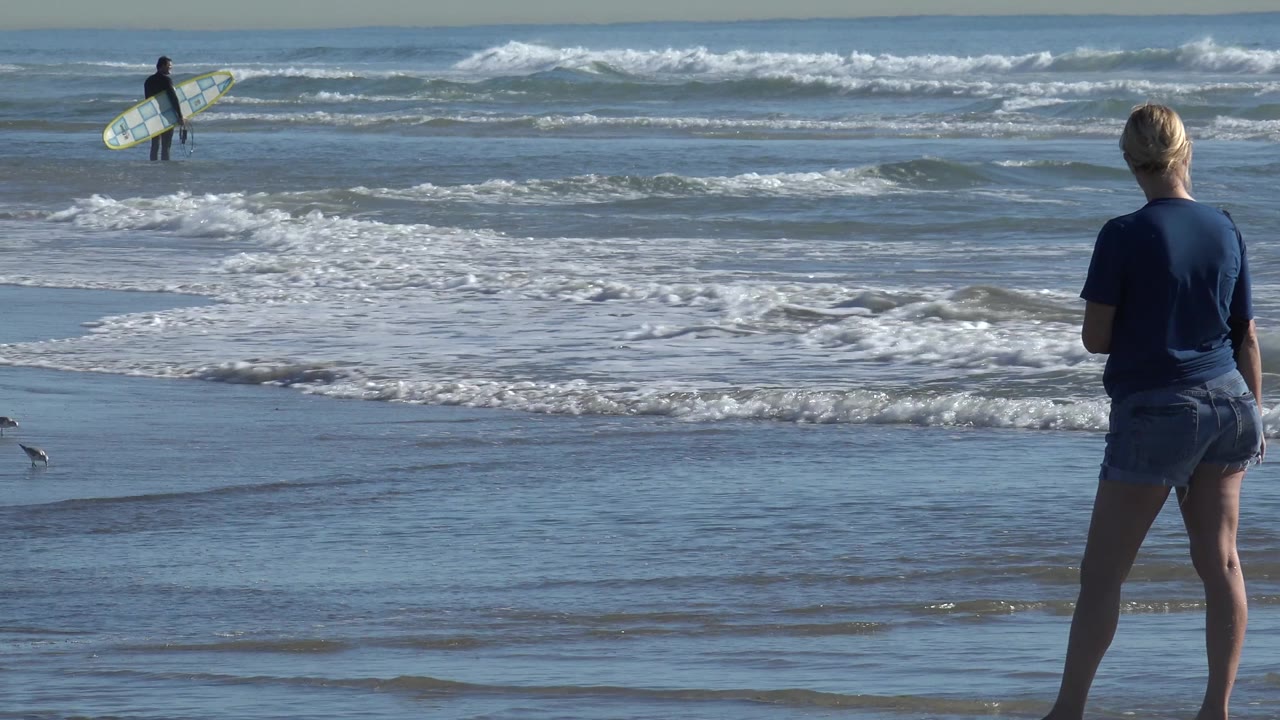 At Huntington Beach, the foreground woman faces the water as a surfer with a board crosses the shallow surf farther back.