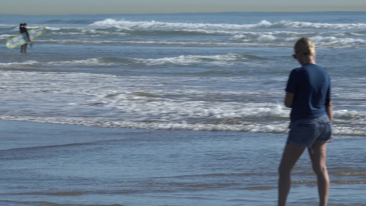 At Huntington Beach, the woman appears near the right side of frame with waves rolling in and a distant surfer moving through the background.