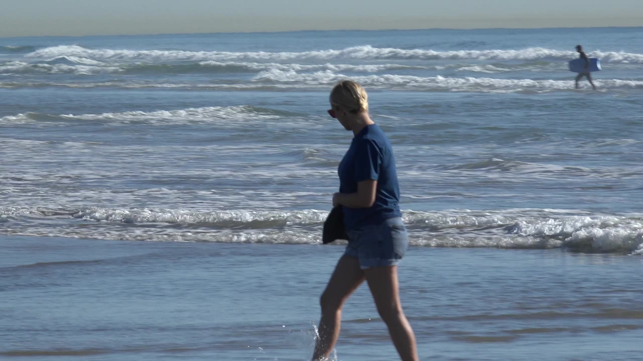 Woman walks barefoot along wet sand with surfers behind her.