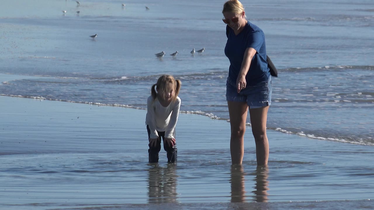 At Huntington Beach, California, a woman in sunglasses stands ankle-deep in reflective water and points down while a child bends near the wet sand.