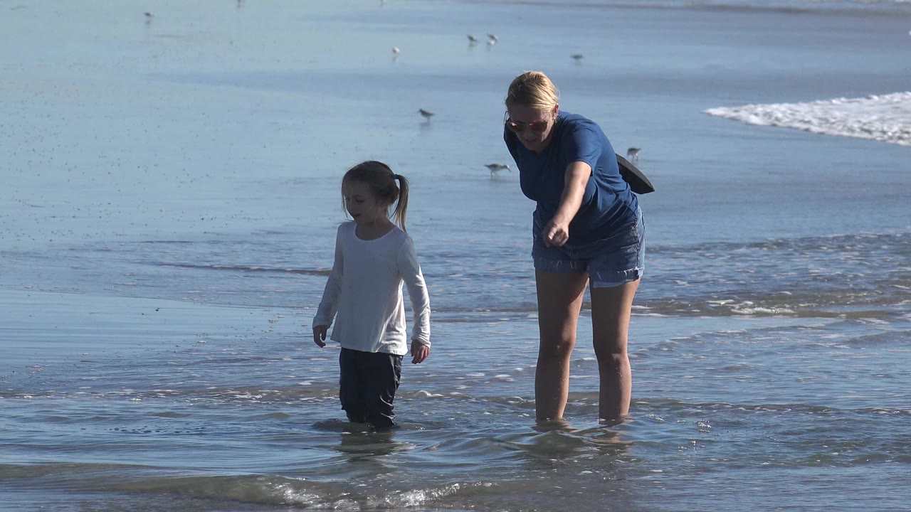 At Huntington Beach, California, a woman bends slightly toward a young girl standing in shallow surf as small waves cross the wet sand behind them.
