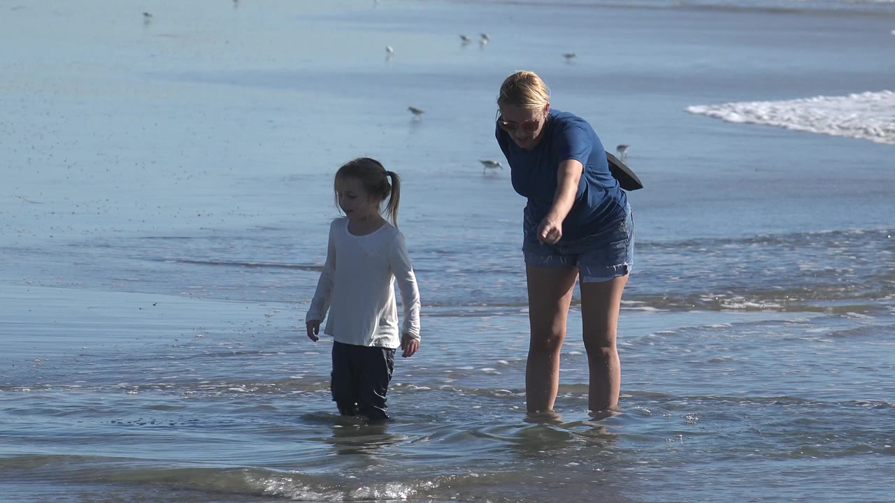 At Huntington Beach, California, the woman points toward the shallow water while the young girl looks ahead, with reflective wet sand and small surf lines behind them.