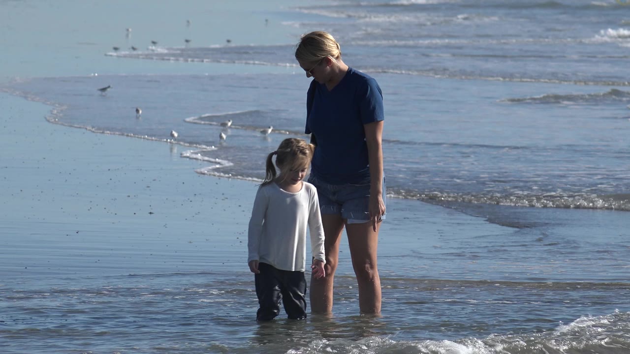 At Huntington Beach, California, the woman looks down toward the child while they stand in shallow surf, with a small wave approaching from the right and gulls behind them.