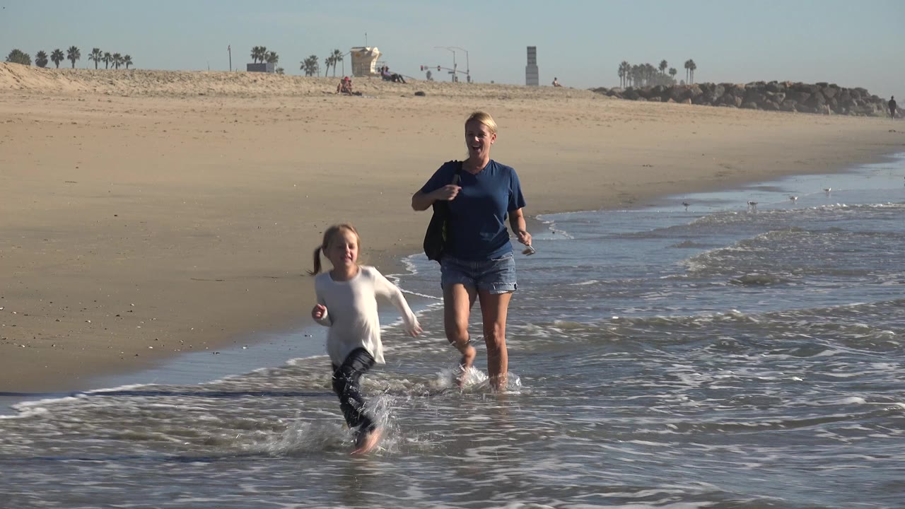 An adult woman and young girl move through shallow surf at Huntington Beach, California, with wet sand in front and a wide beach horizon behind them.