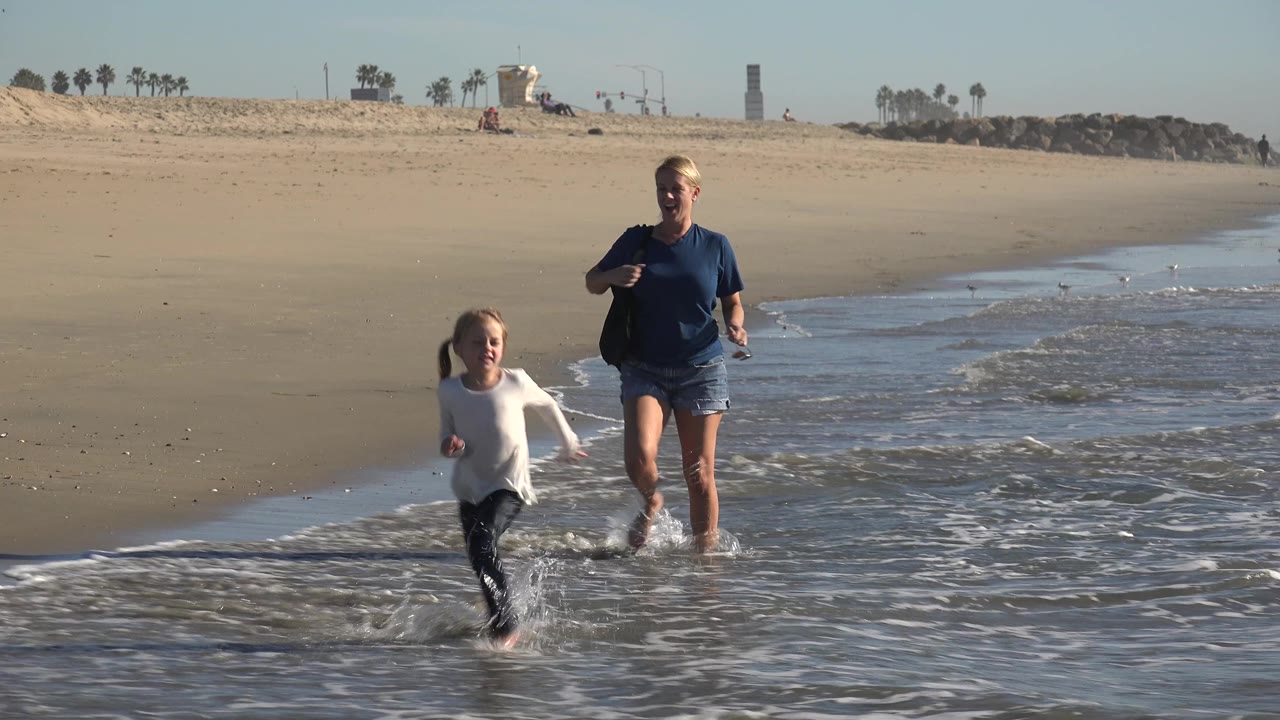 The young girl runs ahead through white foam while the adult woman follows in the shallow water at Huntington Beach, California.