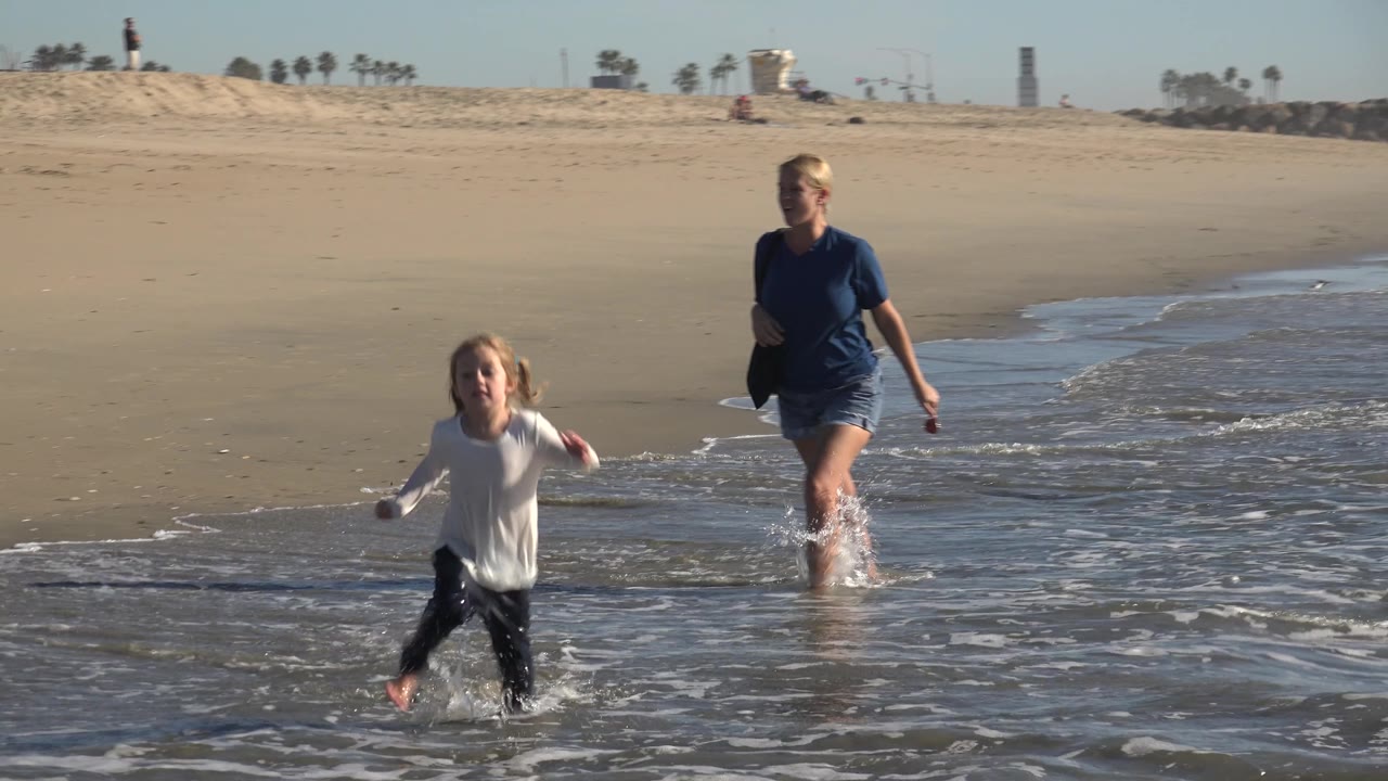 The girl crosses the shallow water in the foreground while the adult woman trails behind near the wave line on the open Huntington Beach shoreline.