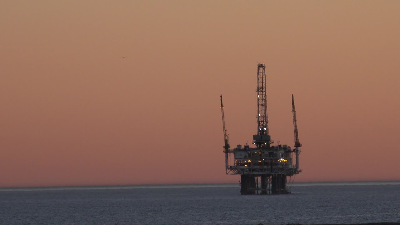 Offshore platform off Huntington Beach sits left of center on a dark ocean horizon under orange dusk light, with no visible people.
