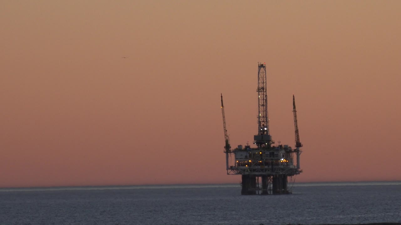 The Huntington Beach offshore platform is framed near center with small lights visible, a flat ocean band below, orange sky above, and no visible people.