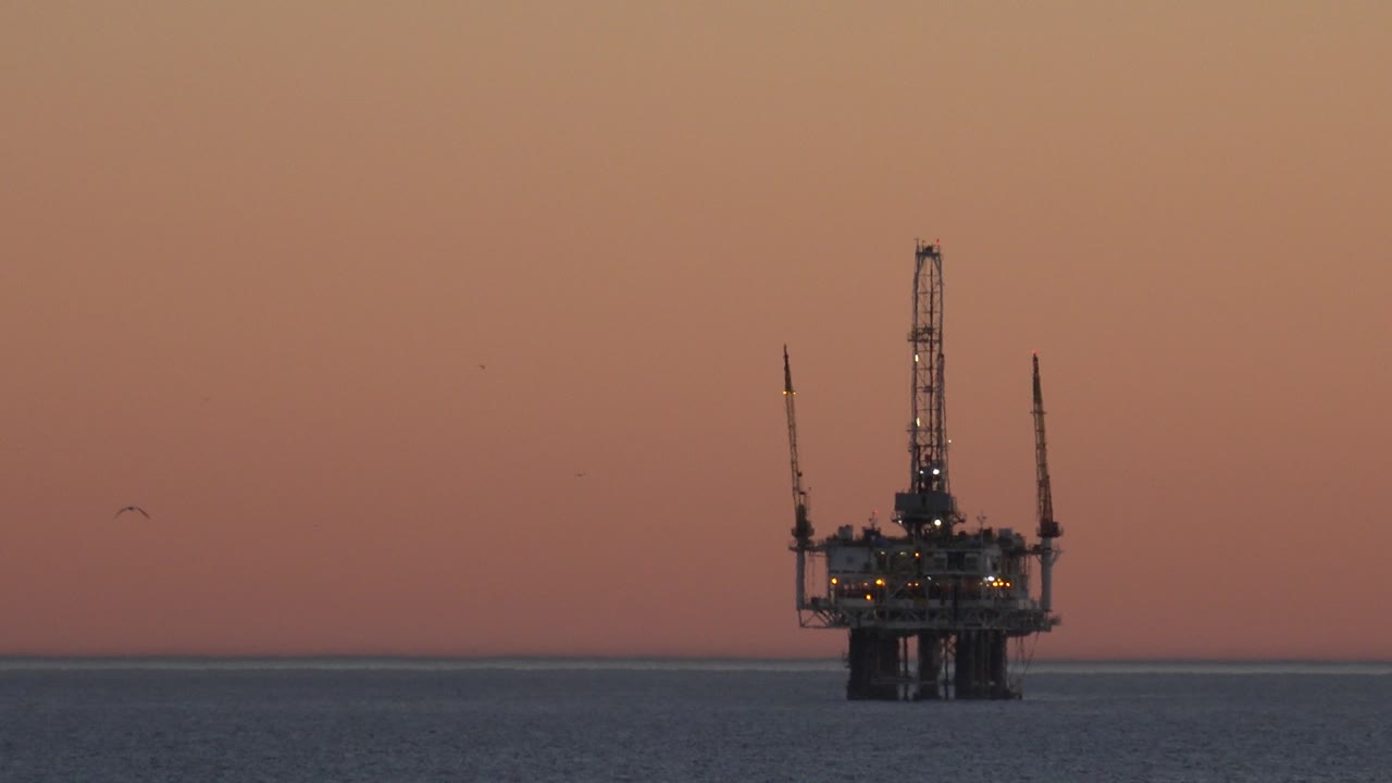 An offshore platform stands near the Huntington Beach ocean horizon under a peach sunset sky, with calm water below and no visible people.