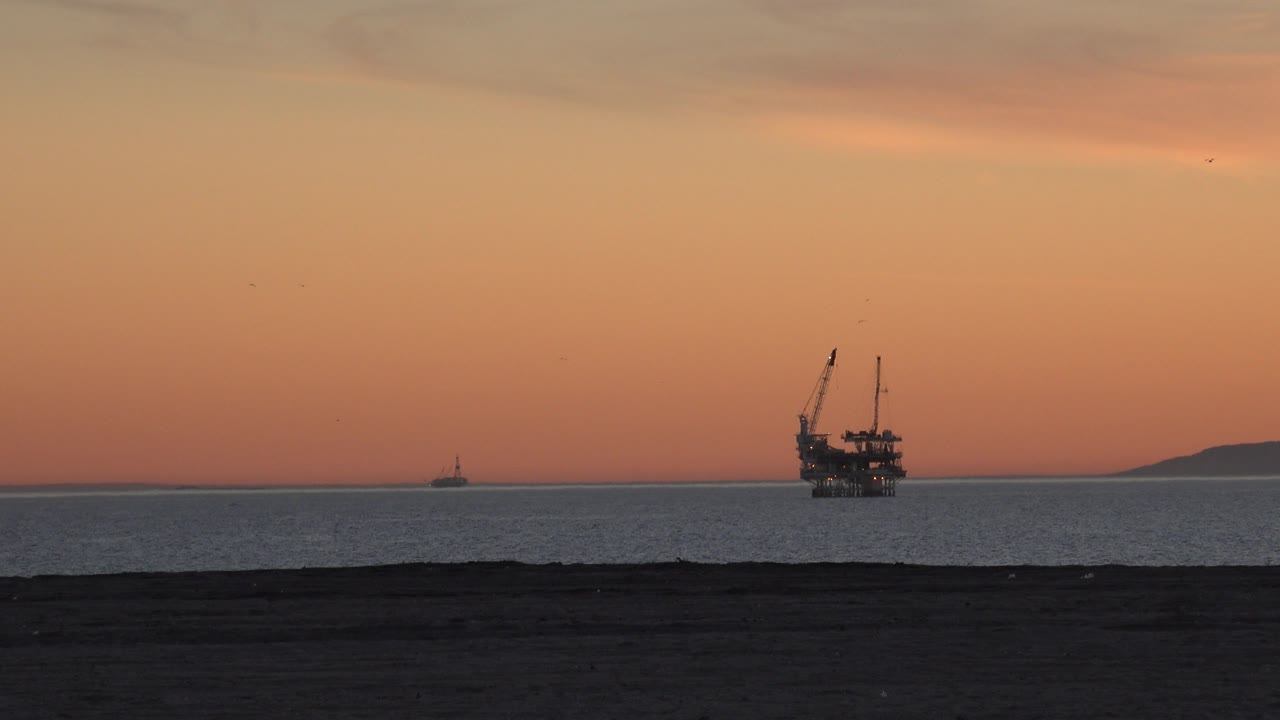 Huntington Beach dusk view with dark beach in the foreground, calm sea, an offshore oil platform near the horizon, low coastline at right, orange sky, and no visible people.