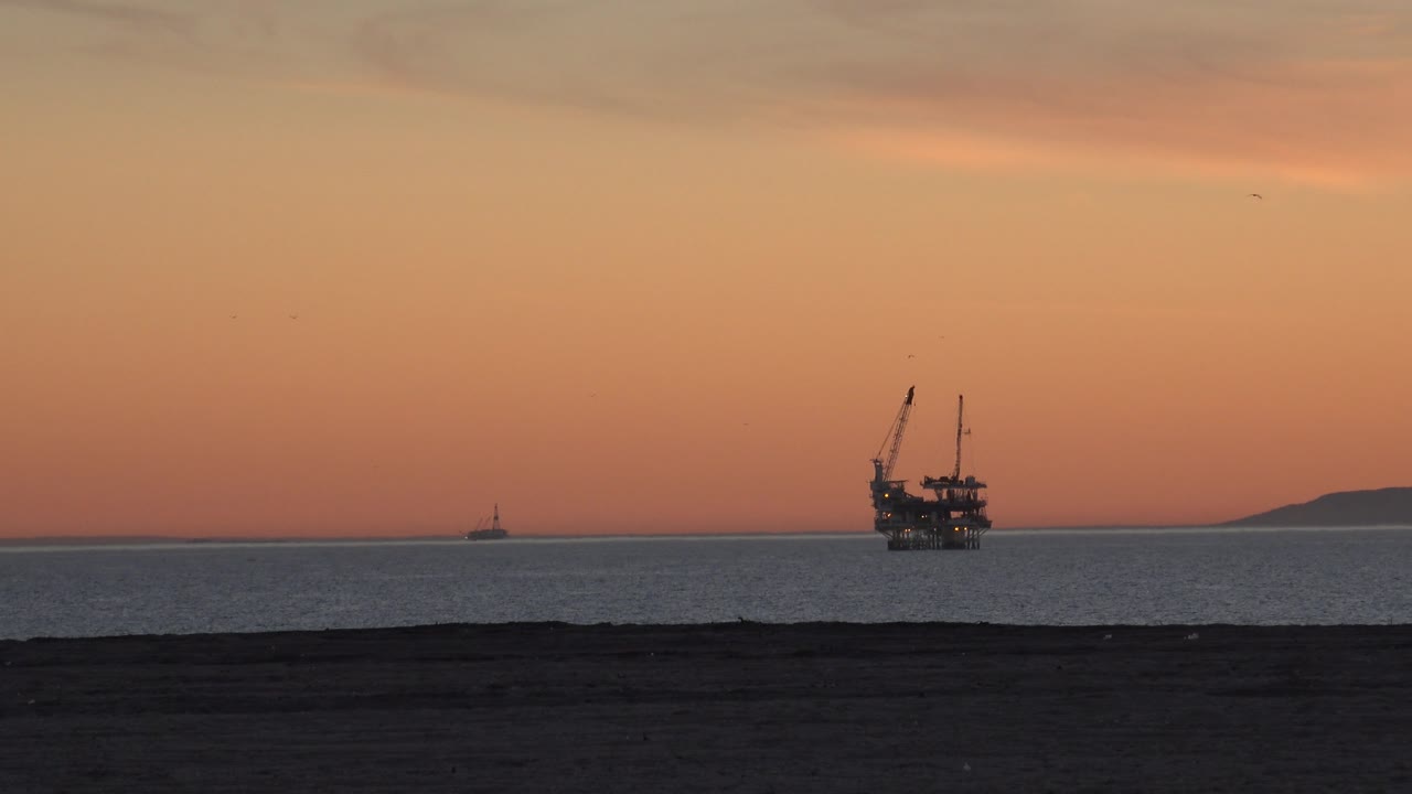 Evening sea view with silhouetted oil platform, distant coastline, and dark sand.