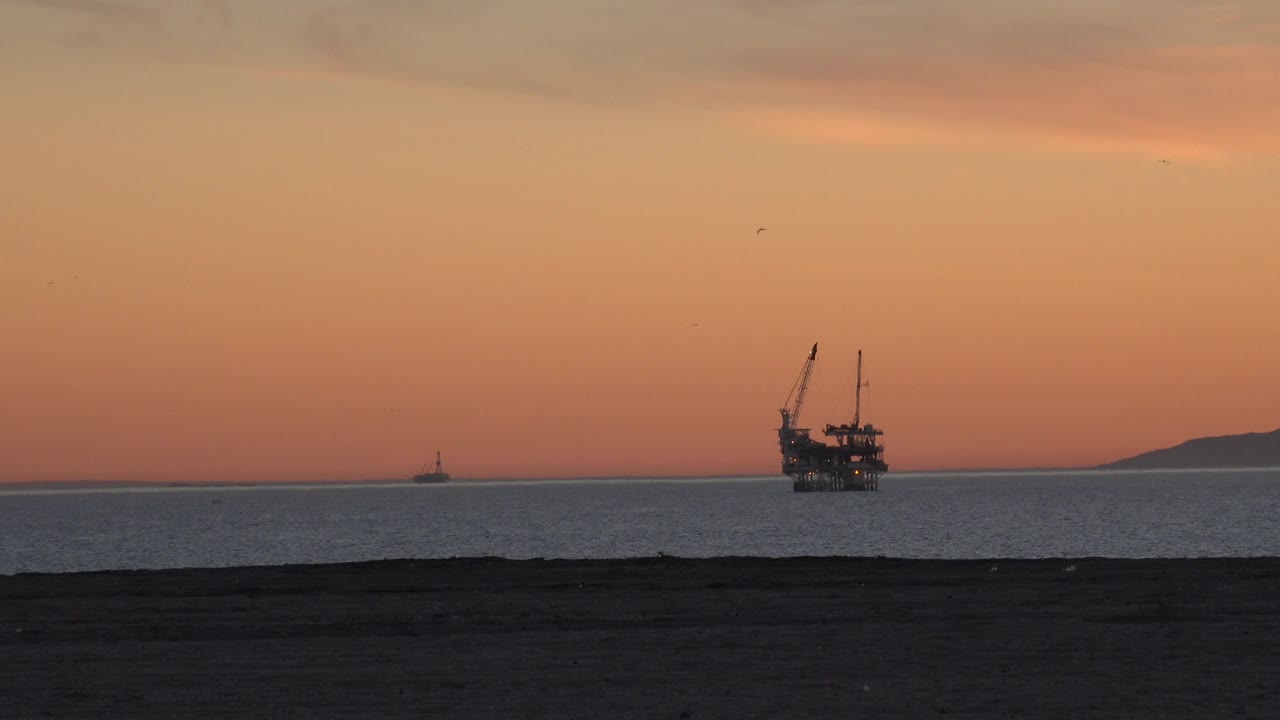 Huntington Beach ocean view at 0:00 with a dark beach foreground, orange sky, an offshore platform near the horizon, distant land at right, and no visible people.