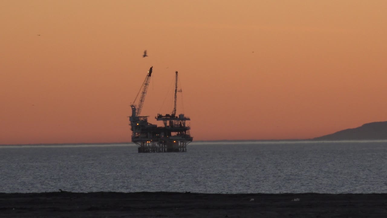 Offshore petroleum platform near Huntington Beach sits left of center beyond calm ocean water, with an orange sky, dark beach foreground, distant land at right, and no visible people.