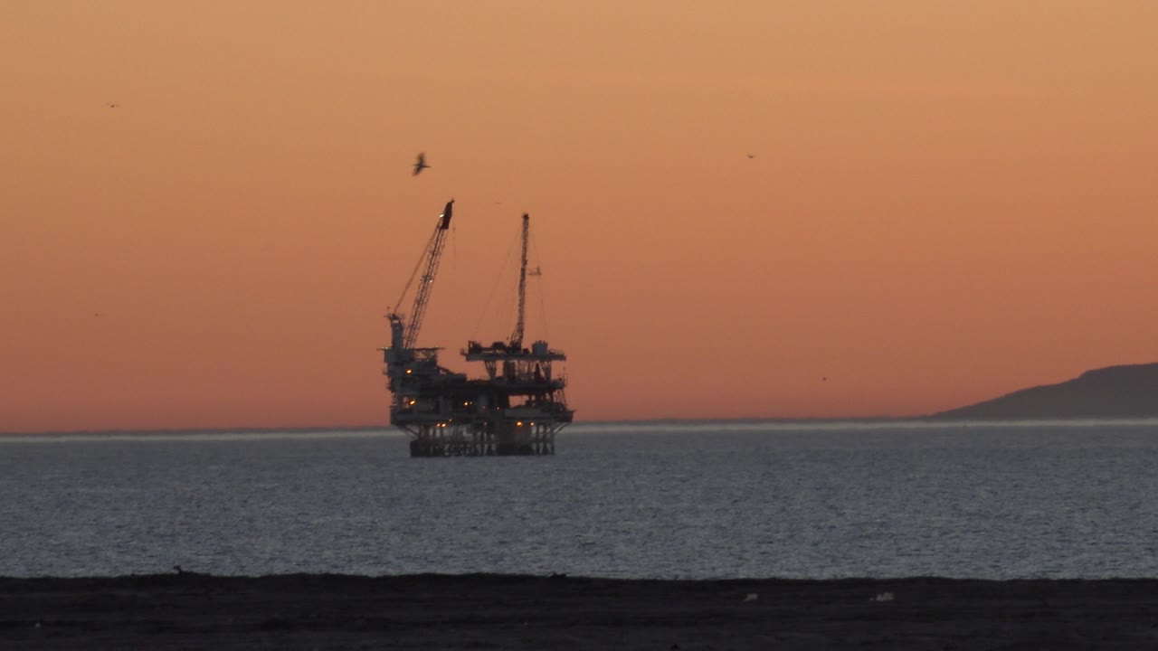 At 0:01, the Huntington Beach ocean view holds the petroleum platform and cranes against orange afternoon light, with a low dark shoreline band and no visible people.
