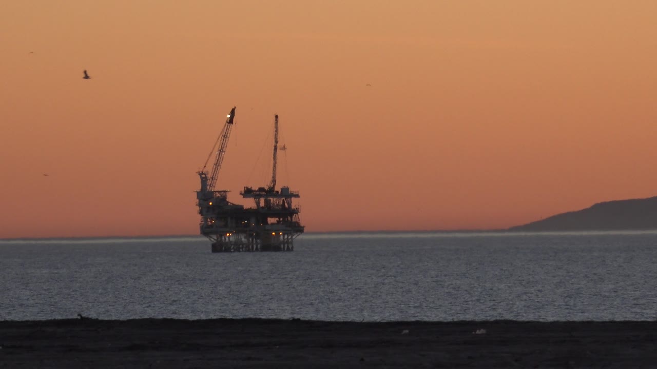 Oil platform lights and cranes visible across the water at dusk-toned light.