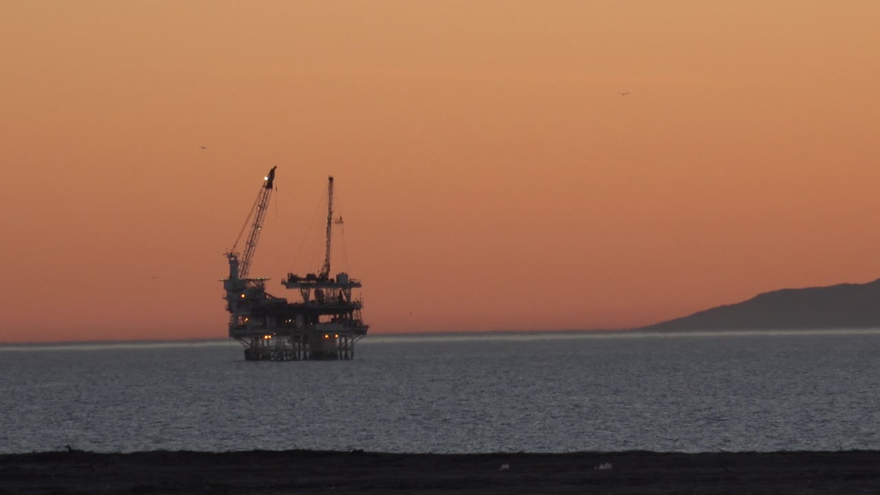 Offshore platform off Huntington Beach at sunset, with a dark shoreline foreground, calm ocean, distant landform on the right horizon, and no visible people.