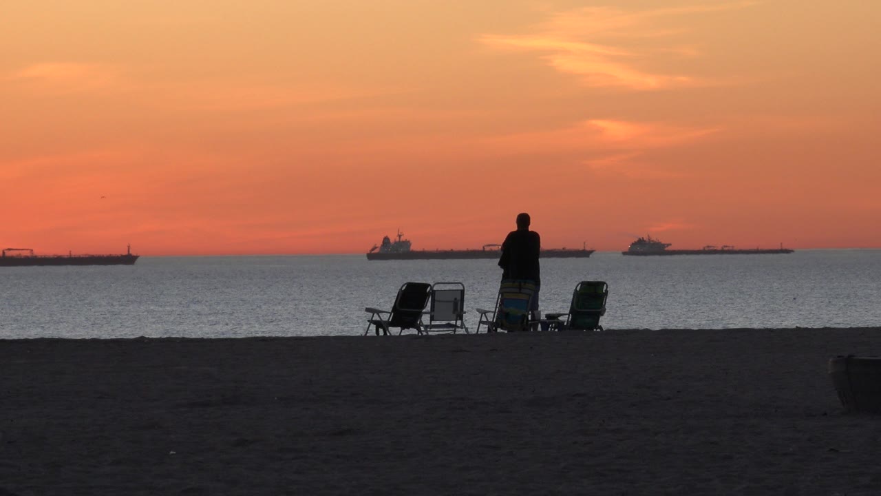 At Huntington Beach, a silhouetted standing person and a seated companion sit near folding chairs on dark sand as orange sunset light spreads over the ocean and distant ships.