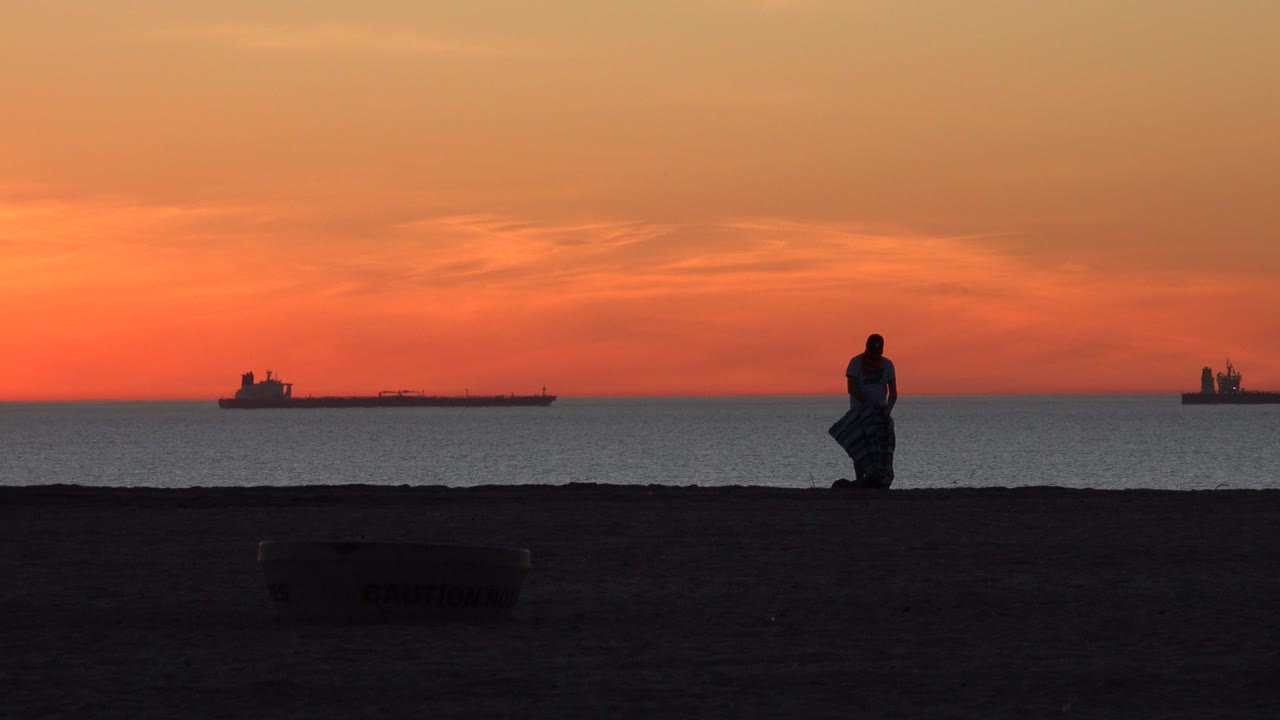 Silhouetted surfer on a dark beach at orange sunset with ships offshore.