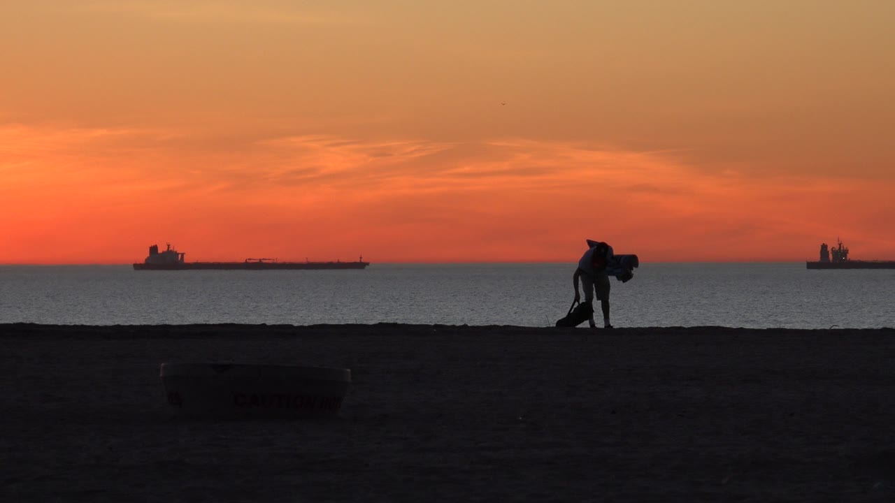 At Huntington Beach, California, a silhouetted person bends near the shoreline while orange sunset light spreads over the ocean and distant ships.