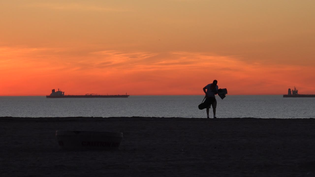 Silhouetted person standing with beach gear near the ocean at sunset.