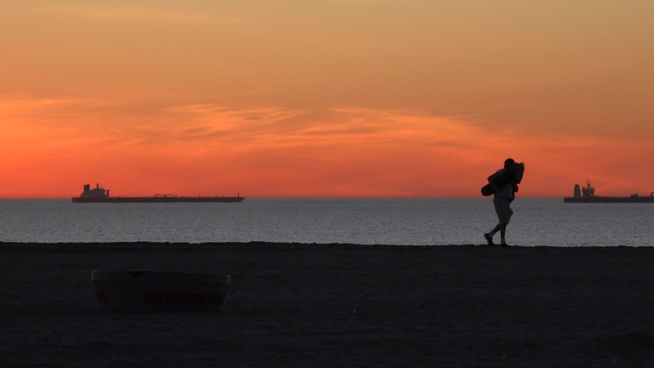 Orange sunset over ocean with ships and a silhouetted beach walker.