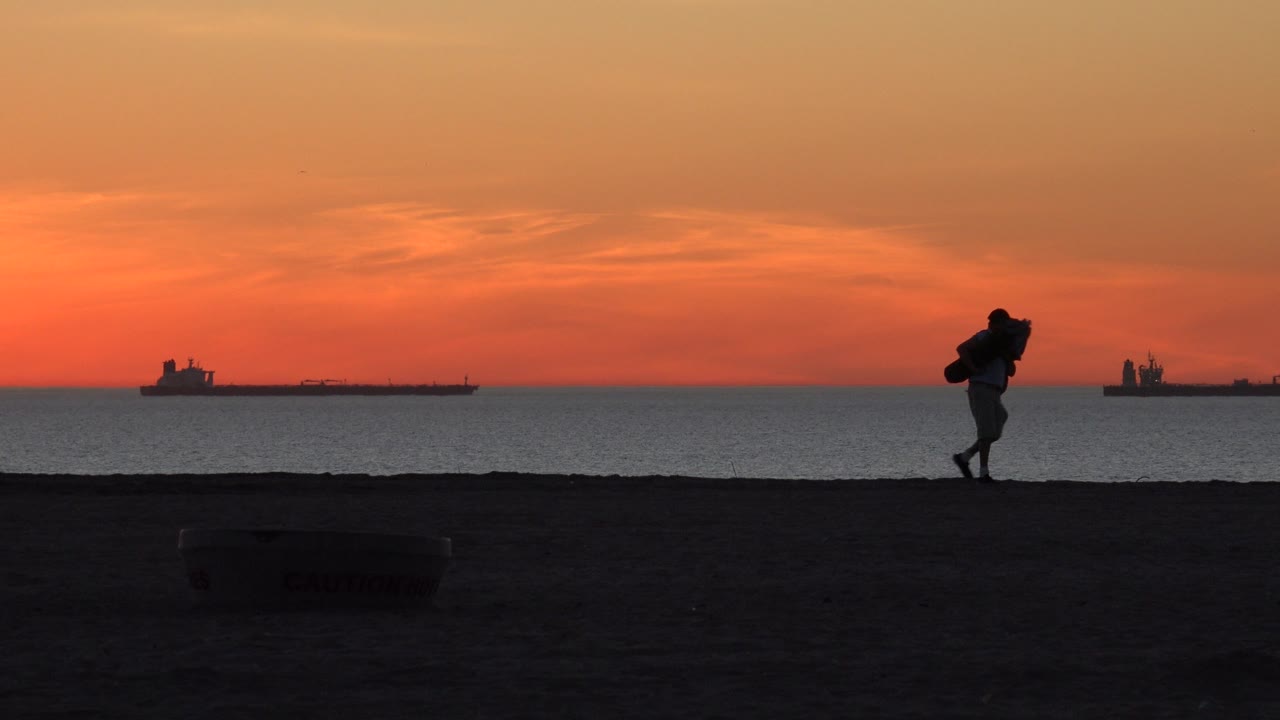 At Huntington Beach, California, the orange sky fills the upper frame as a dark beach walker moves near the right side and ships remain on the horizon.