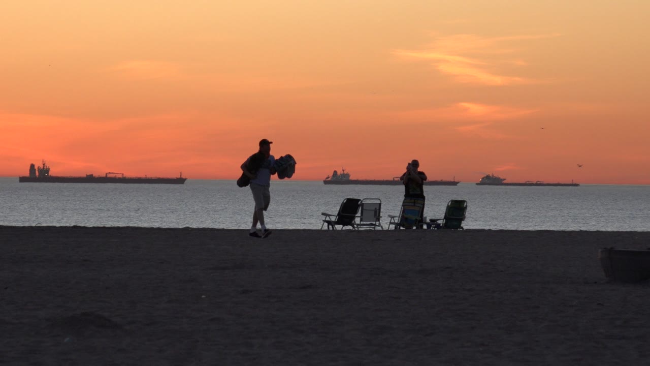 At Huntington Beach, California, a surfer carries a board across the dark sand while another person stands beside folding chairs and ships sit on the orange horizon.