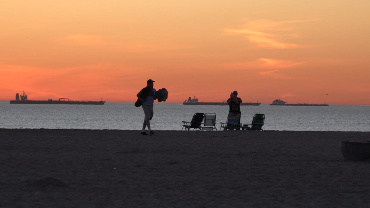 At Huntington Beach, California, the surfer continues across the sand with the board in hand as folding chairs and a second beachgoer remain near the waterline.