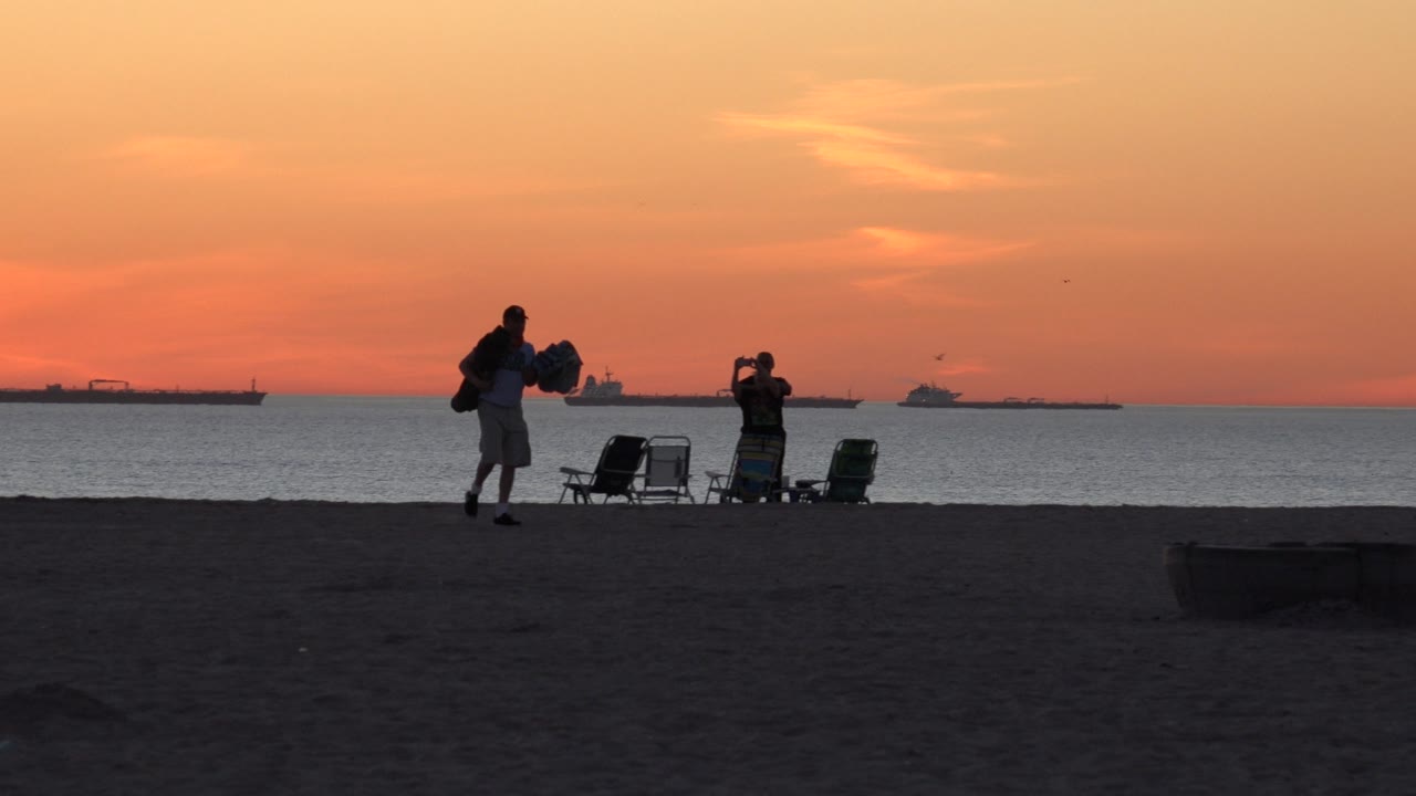 Silhouetted surfer near beach chairs with cargo ships on the sunset horizon.
