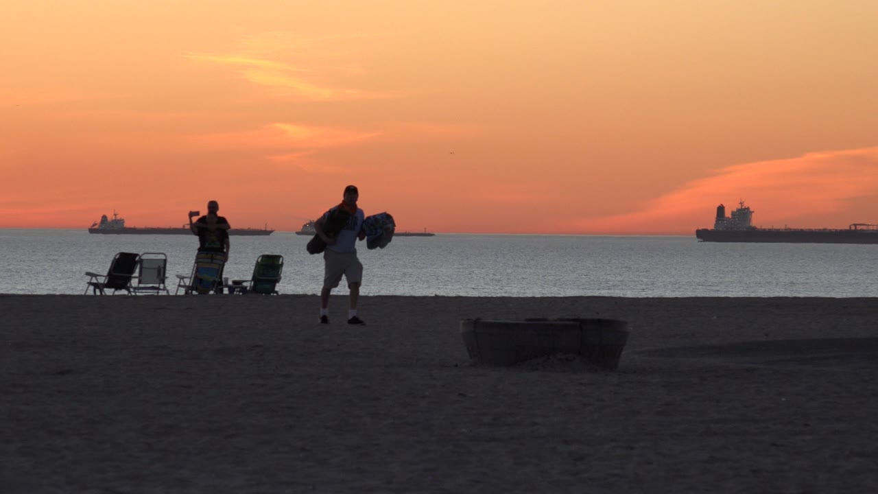 Silhouetted beachgoers, chairs, sand, ocean, and ships under an orange sky.