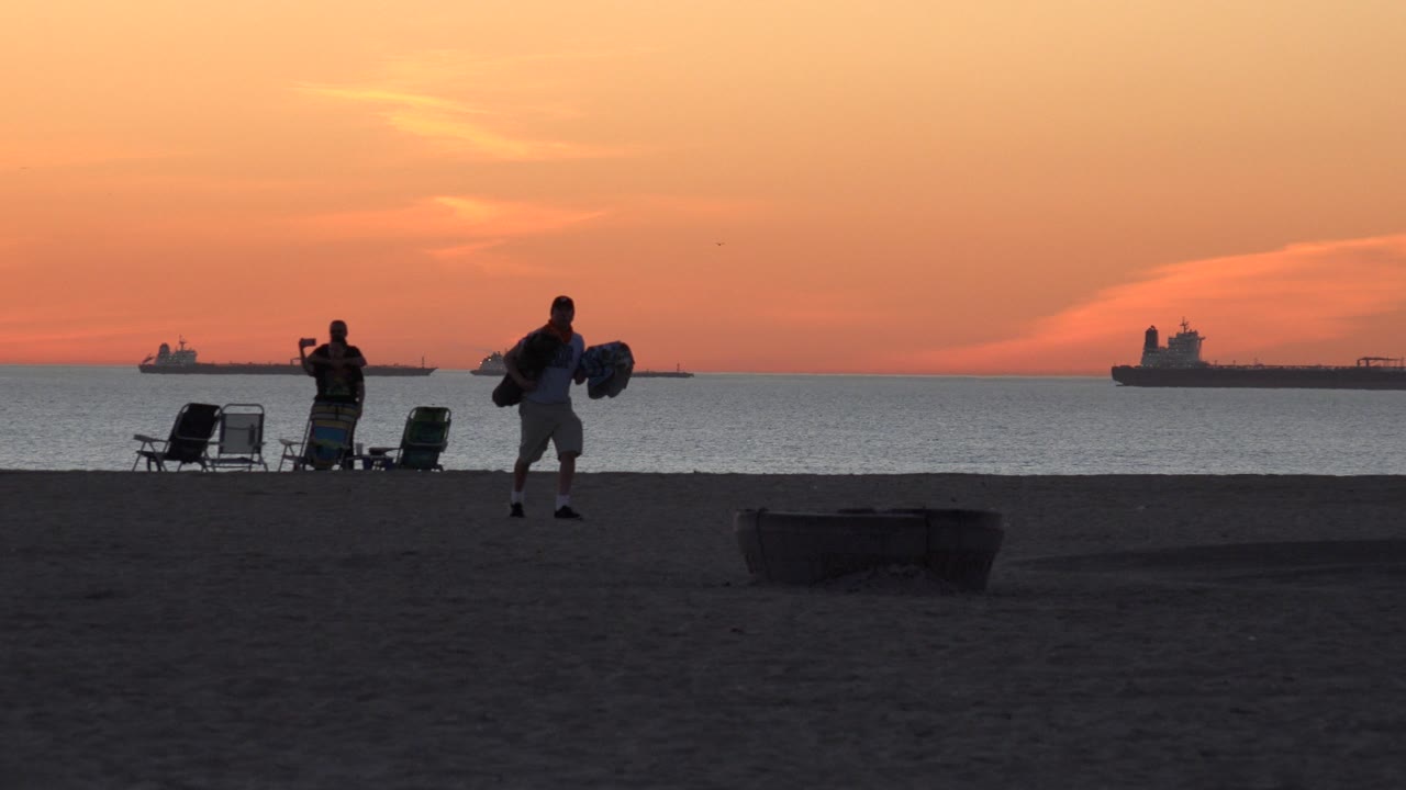 At Huntington Beach, two silhouetted beachgoers are separated across the sand, with chairs clustered near the waterline and cargo ships beyond them.