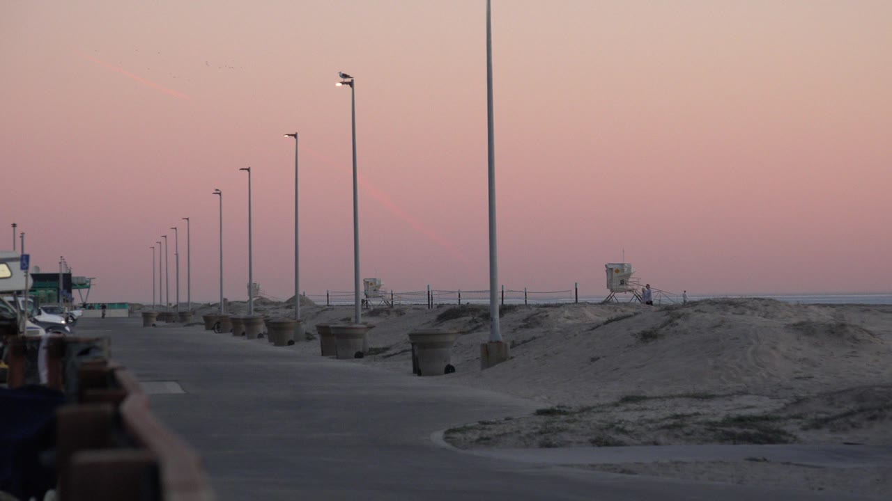 Opening frame of Huntington Beach with a paved beach path, row of lamps, sand berms, distant lifeguard tower, pink dusk sky, and no clearly visible people.