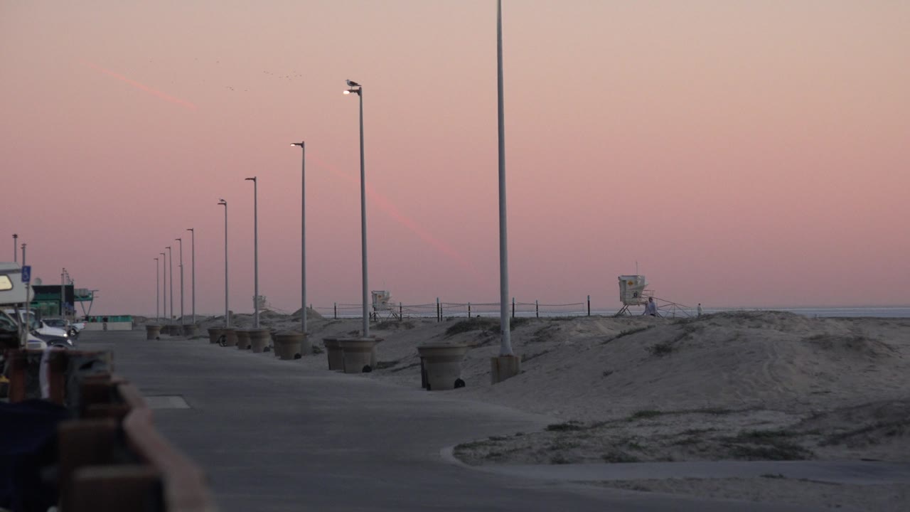 A Huntington Beach dusk frame with the path running left of sand berms, tall lamps spaced into the distance, the lifeguard tower near the horizon, and no clearly visible people.