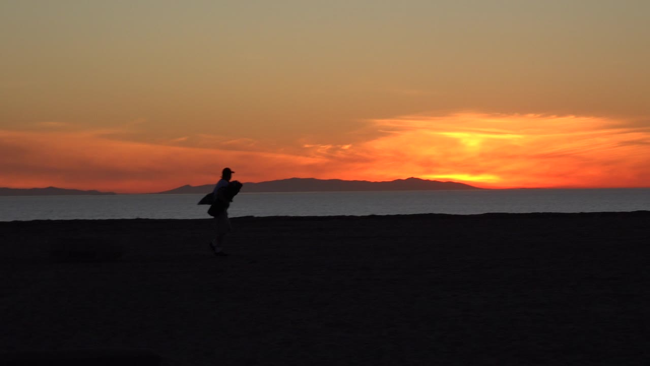Silhouetted backpacked walkers on a dark beach at sunset.