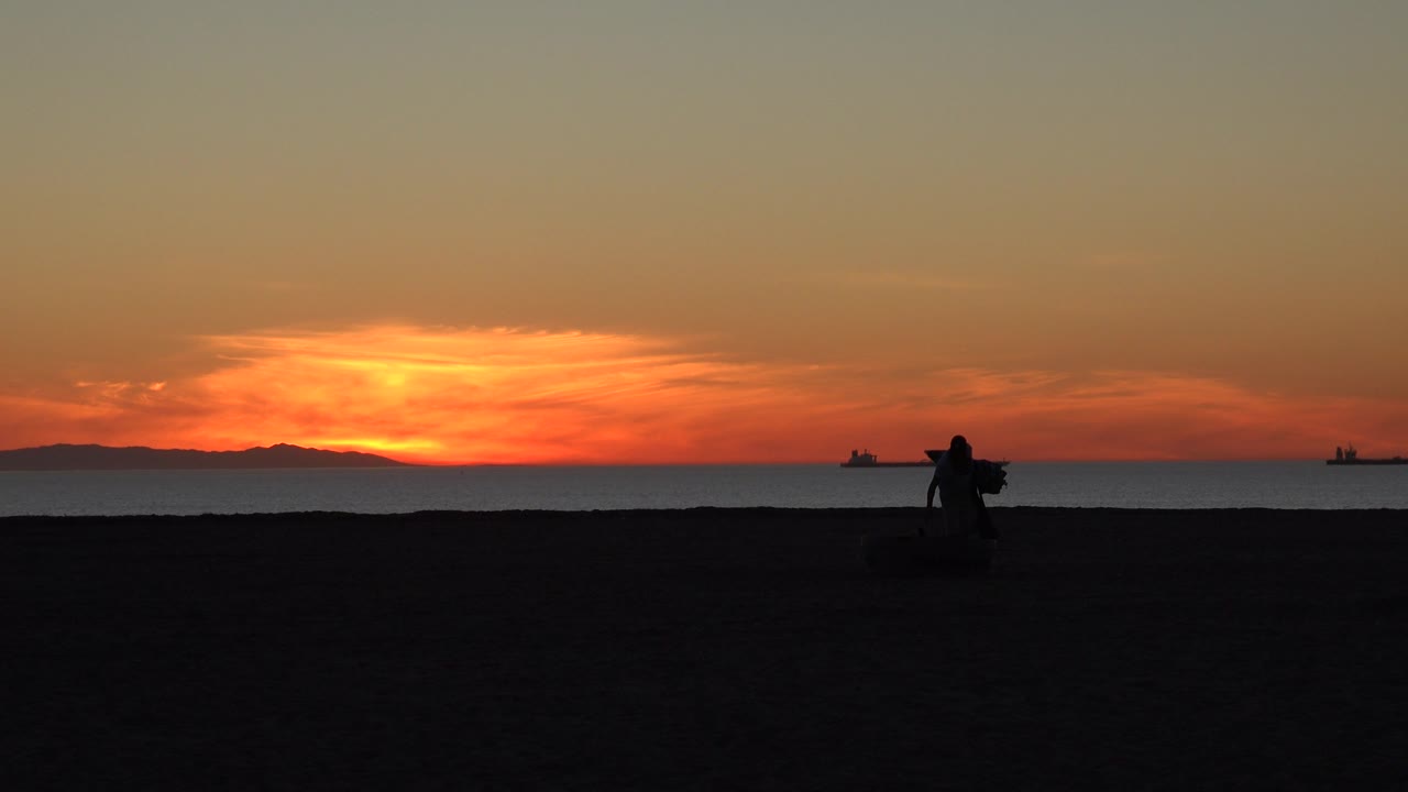 A dark figure remains near the Huntington Beach shoreline as the sunset glow spreads across low clouds above the ocean and distant land.