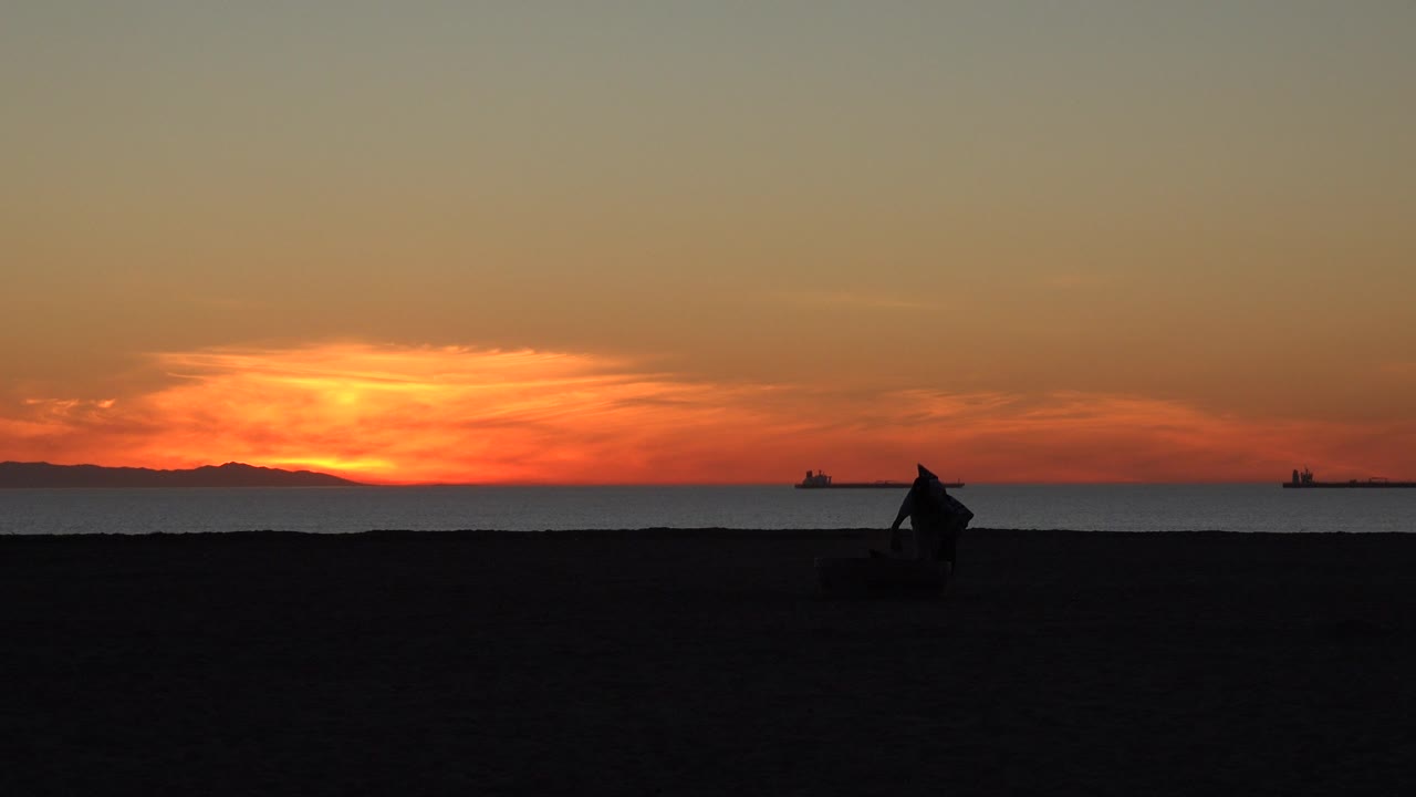 The Huntington Beach shoreline remains dark in the foreground while the silhouetted cleaner is closer to center, with ships and a low orange sunset band behind him.