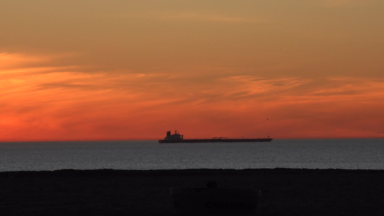 Huntington Beach ocean sunset with the cargo ship still left of center, a brighter orange cloud band above it, dark water below, and no visible people.