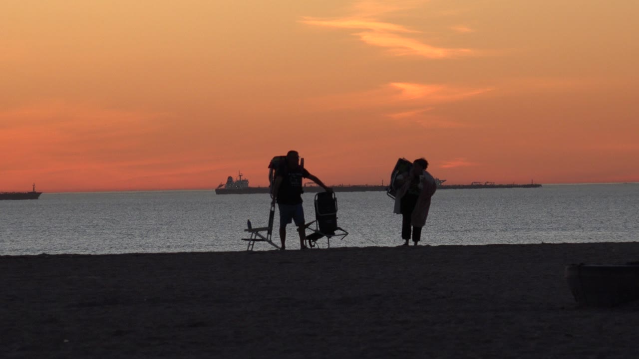 Two silhouetted adults with beach chairs on dark sand at sunset.