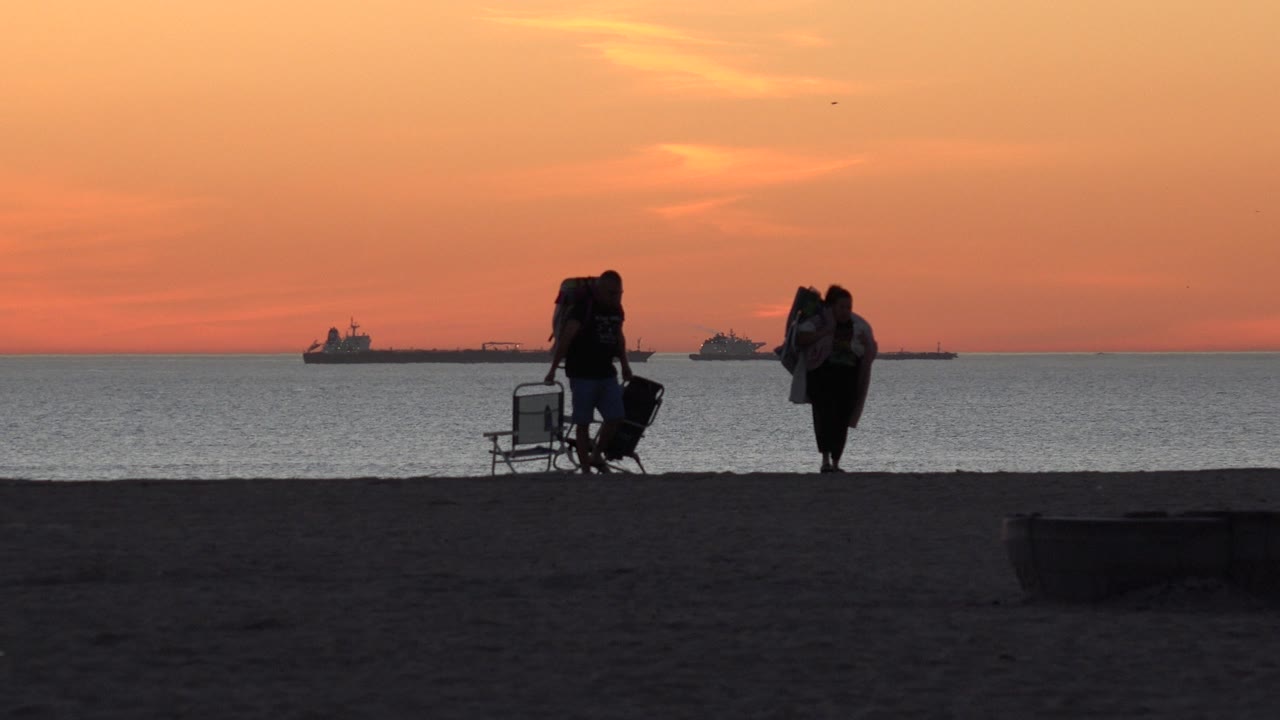 Two silhouetted people stand near the Huntington Beach shoreline with folding chairs and bags, dark sand in front, ships on the horizon, and orange sunset sky above.