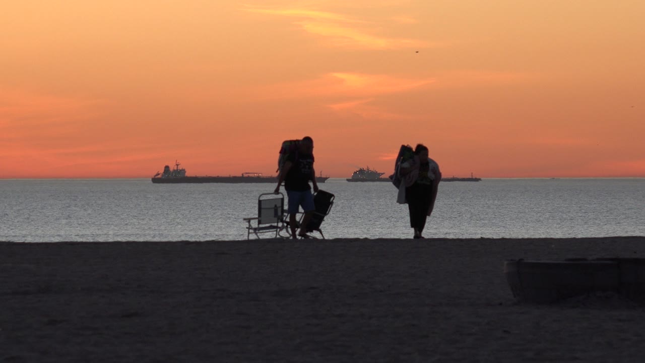 The two beachgoers are slightly farther along the sand at Huntington Beach, carrying chairs and bags against a calm ocean and warm orange horizon.