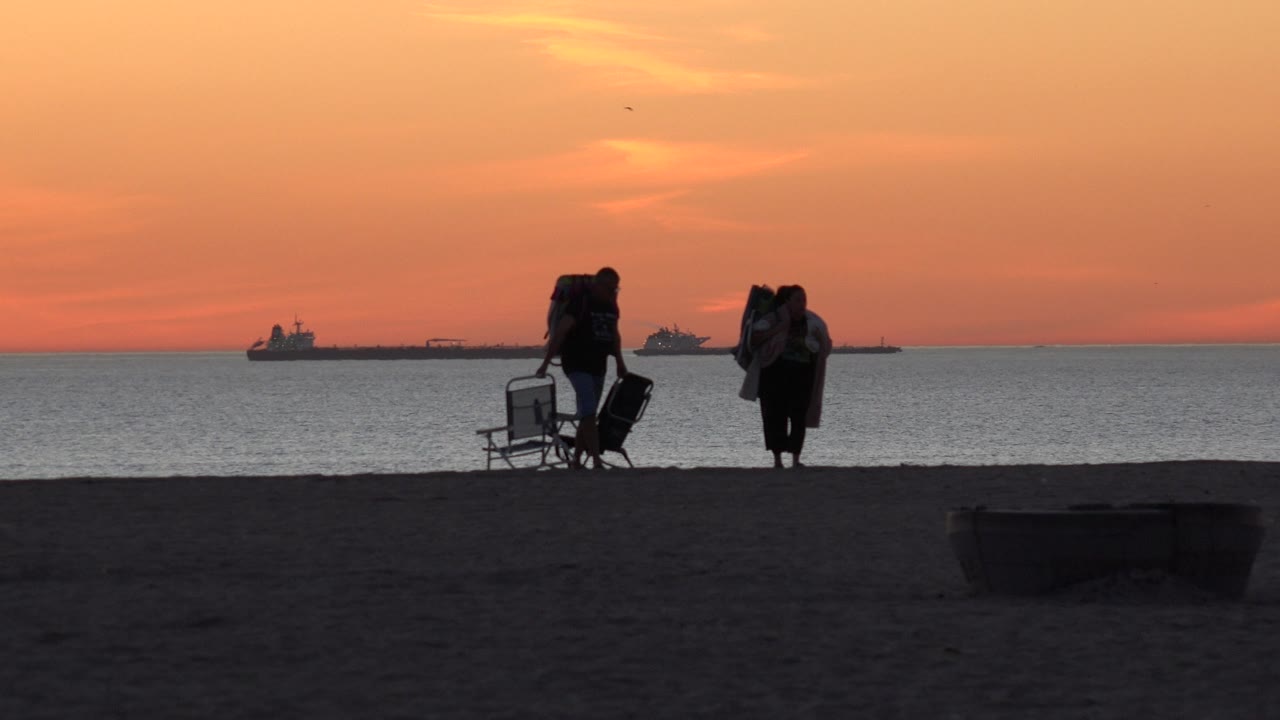 Two silhouetted beachgoers continue across the Huntington Beach sand, with one person beside stacked chairs and the other carrying bags, backed by a flat ocean horizon and sunset sky.